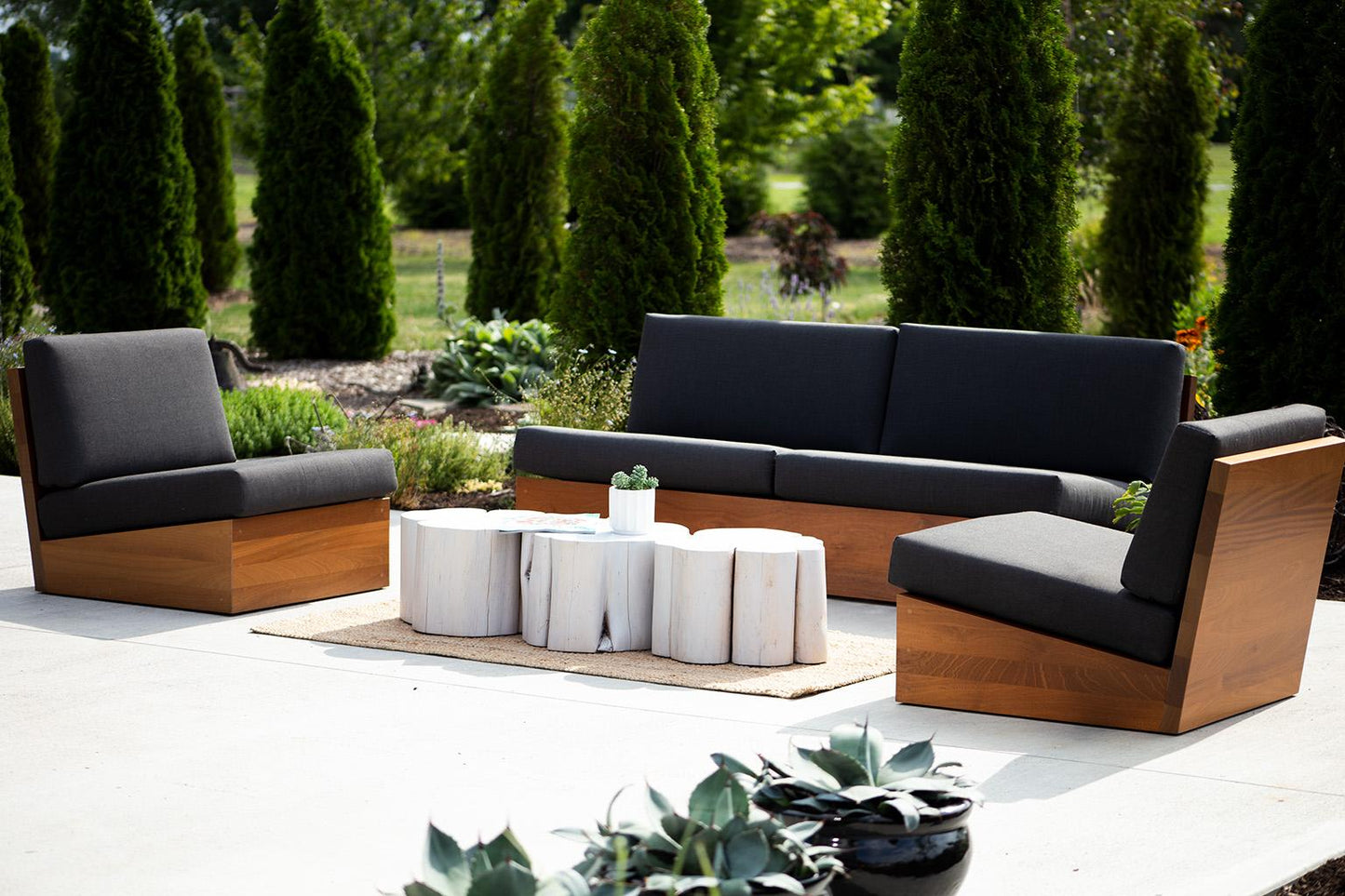 Outdoor patio set with wooden frames and dark cushions on a concrete surface, surrounded by greenery.