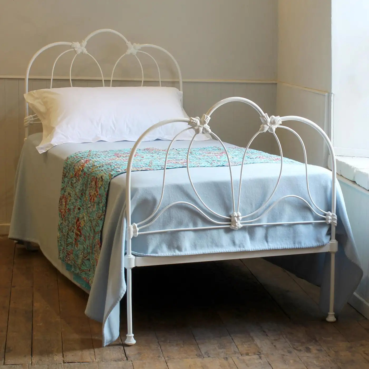 White metal bed with floral bedding in a room with wooden flooring and a window.
