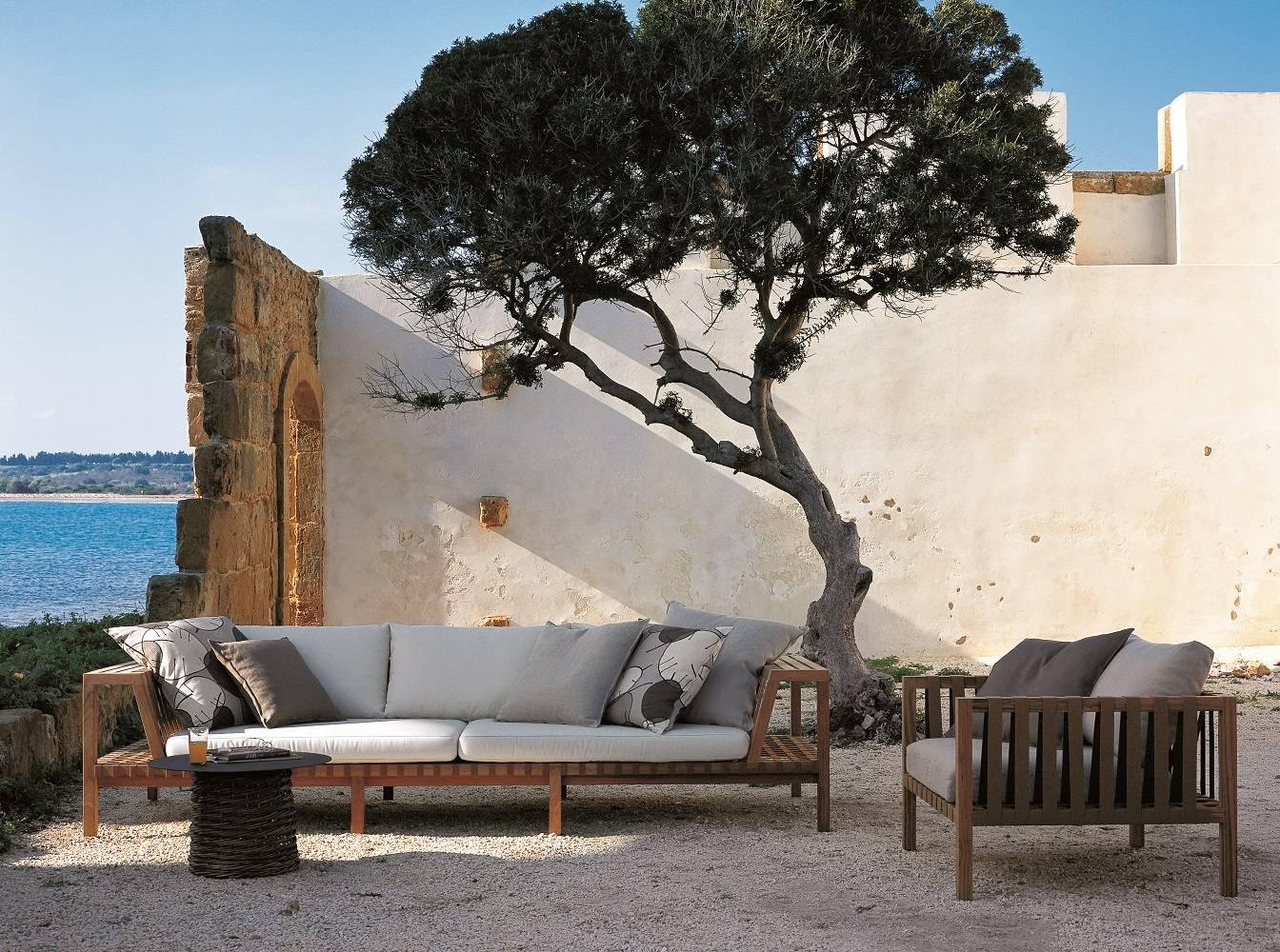 Outdoor seating area with wooden furniture and cushions near a body of water and white wall.