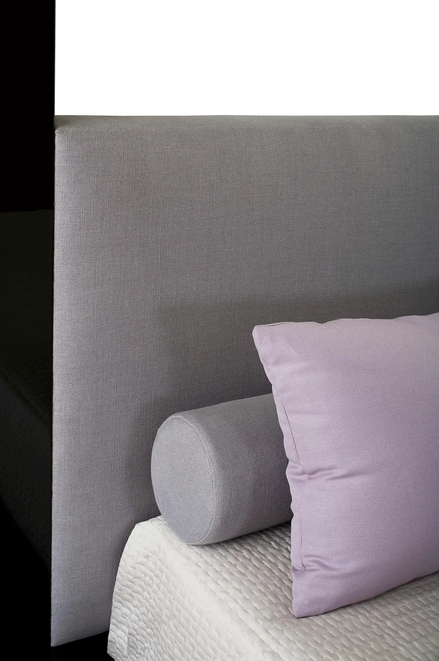 Gray headboard with a cylindrical pillow and a purple pillow on a bed.