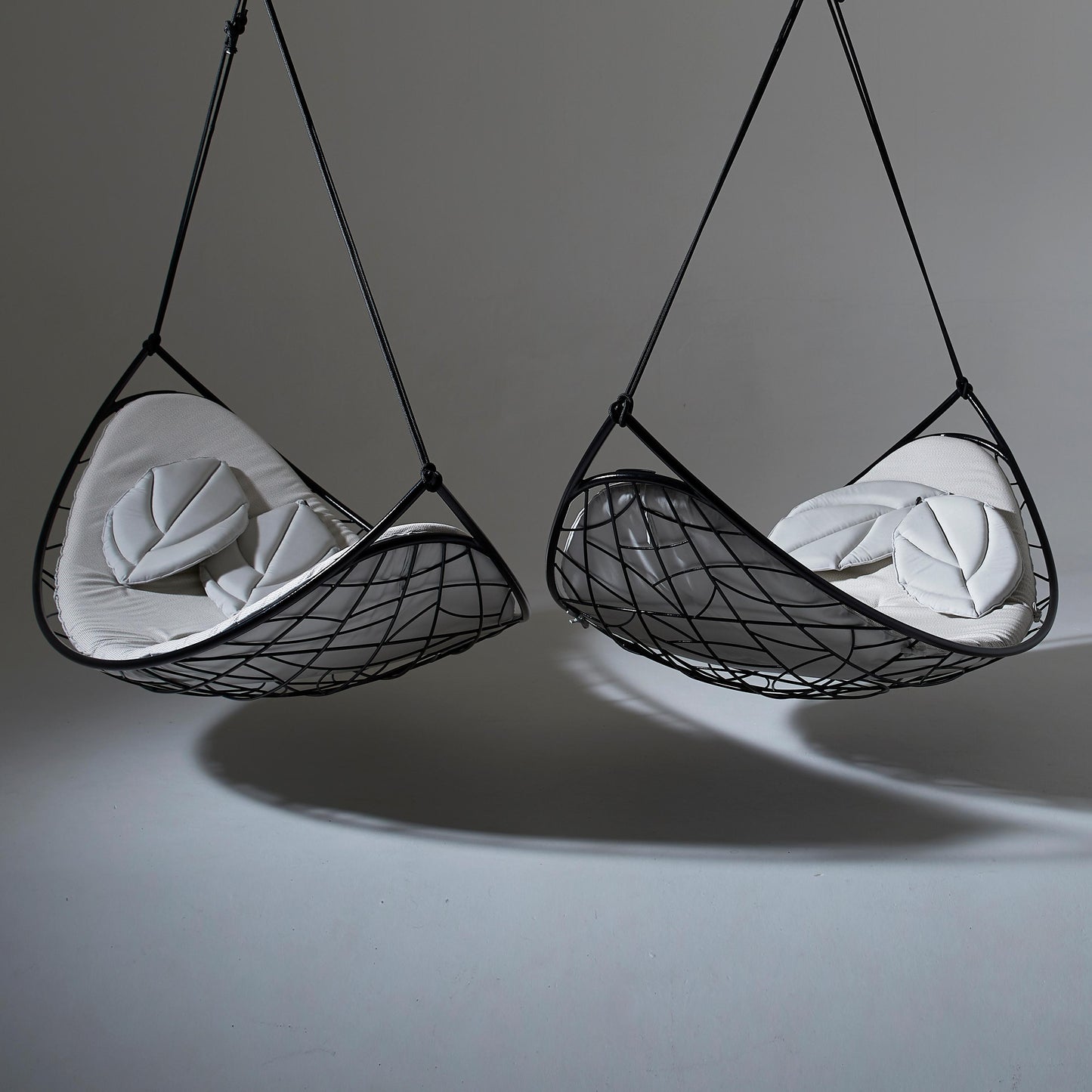 Two black mesh hanging chairs with white cushions on a gray background