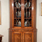 Wooden corner cabinet with glass doors displaying decorative plates against a white wall.