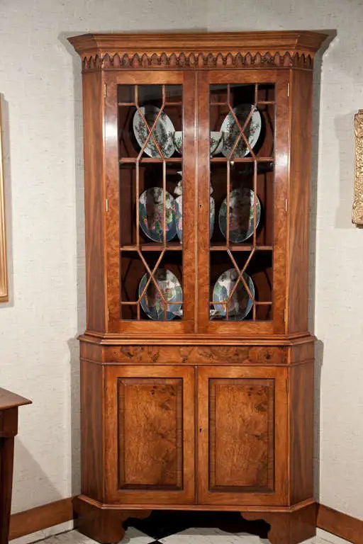 Wooden corner cabinet with glass doors displaying decorative plates against a white wall.