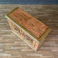 Wooden chest with decorative carvings on a wooden floor against a blue brick wall.