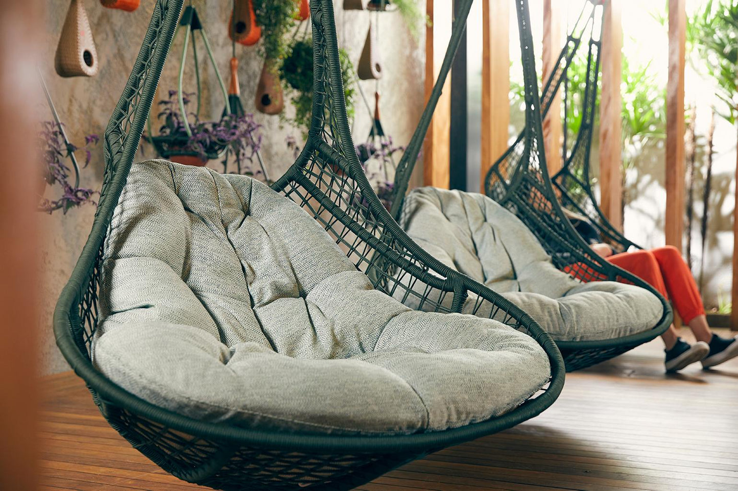 Hanging wicker chairs with cushioned seats in a room with plants and a person sitting on one.