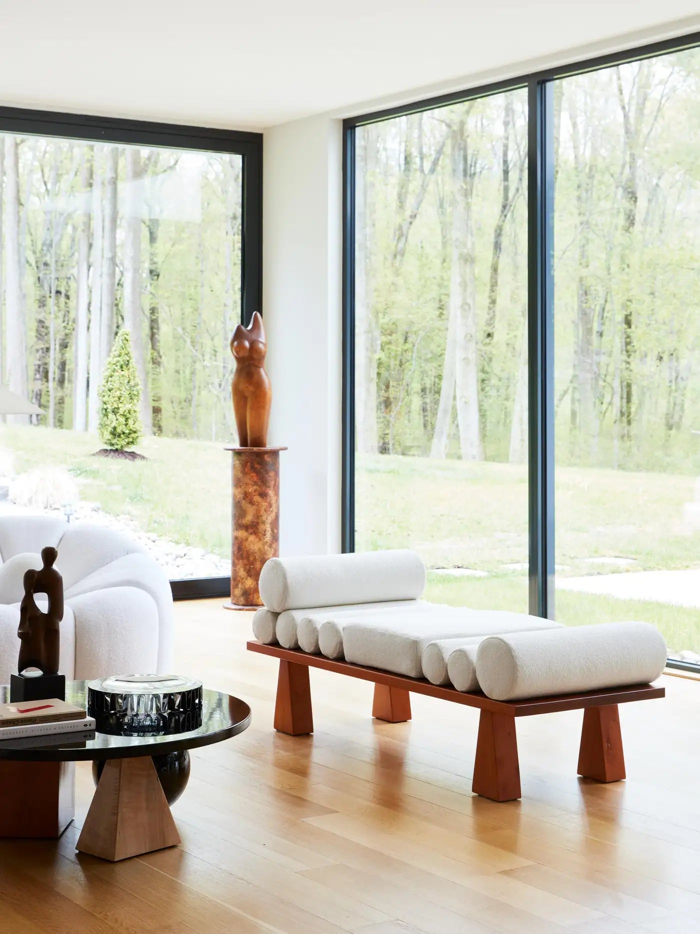 Modern living room with a wooden bench and white cushions, large windows showing a forest view.