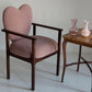 Heart-shaped chair with wooden frame and pink cushion next to a small wooden table with vases.