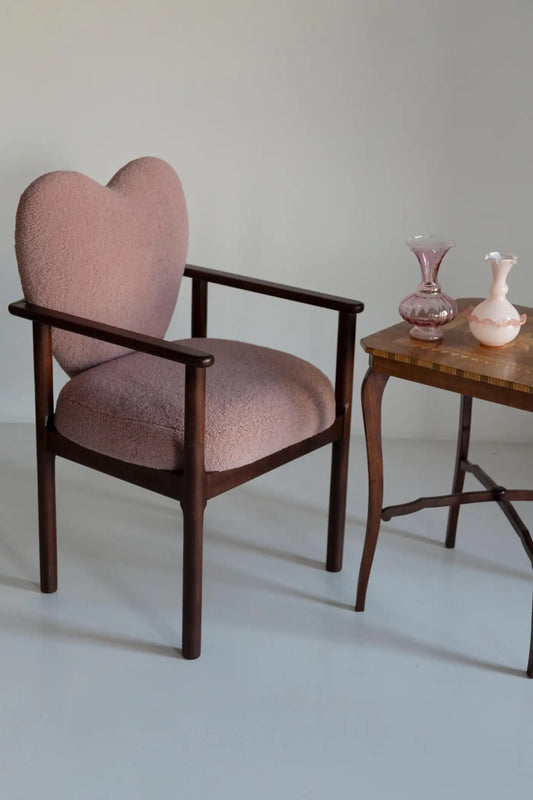 Heart-shaped chair with wooden frame and pink cushion next to a small wooden table with vases.