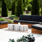 Outdoor patio set with wooden frames and dark cushions on a concrete surface, surrounded by greenery.
