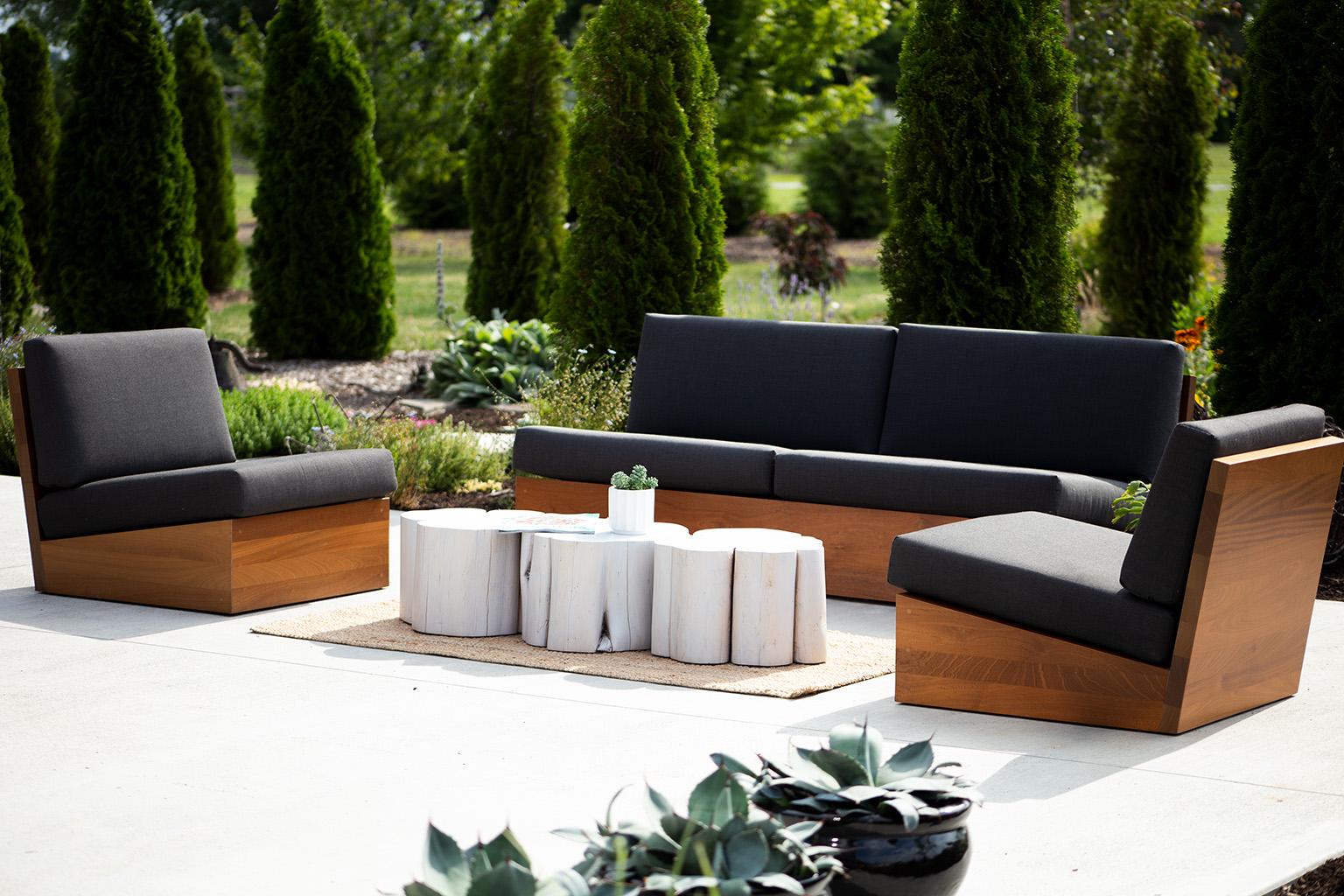 Outdoor patio set with wooden frames and dark cushions on a concrete surface, surrounded by greenery.