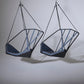 Two geometric hanging chairs with black frames and blue cushions on a gray background