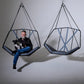 Person sitting in a geometric hanging chair with a dog, against a plain background