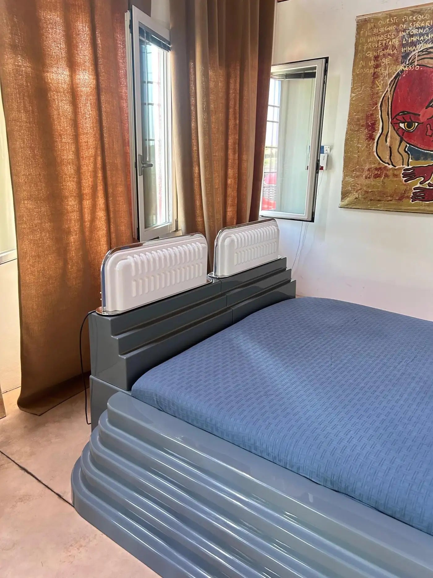 Bed with blue mattress in a room with brown curtains and a painting on the wall.