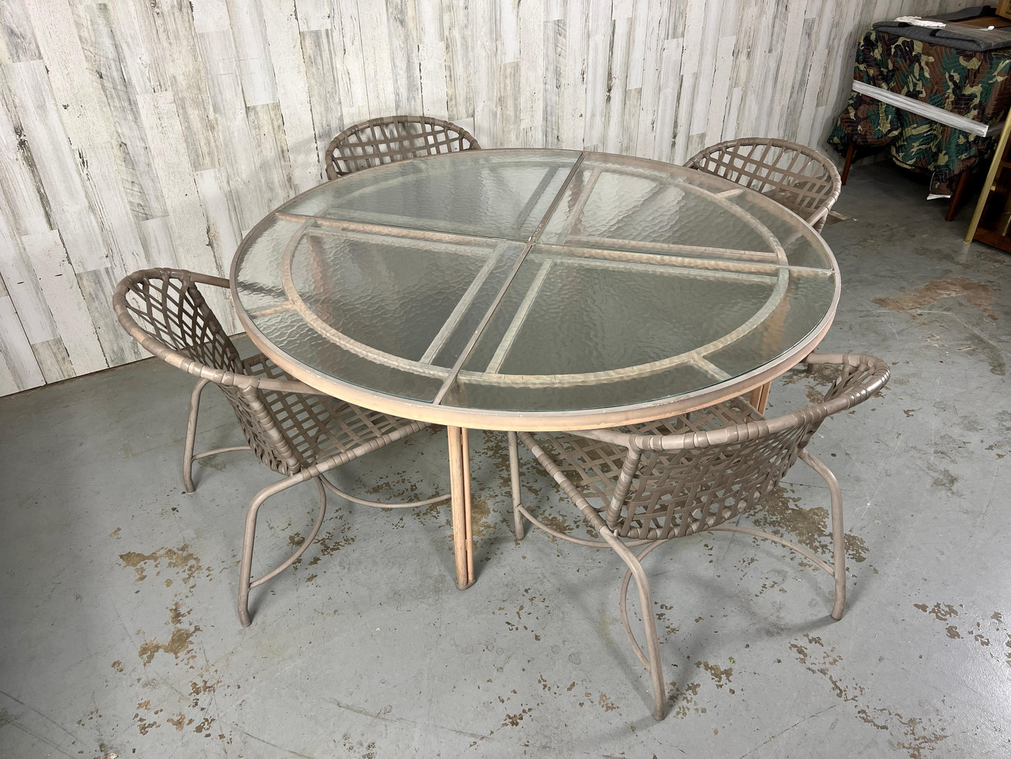 Round glass table with wicker chairs on a concrete floor against a wooden wall.