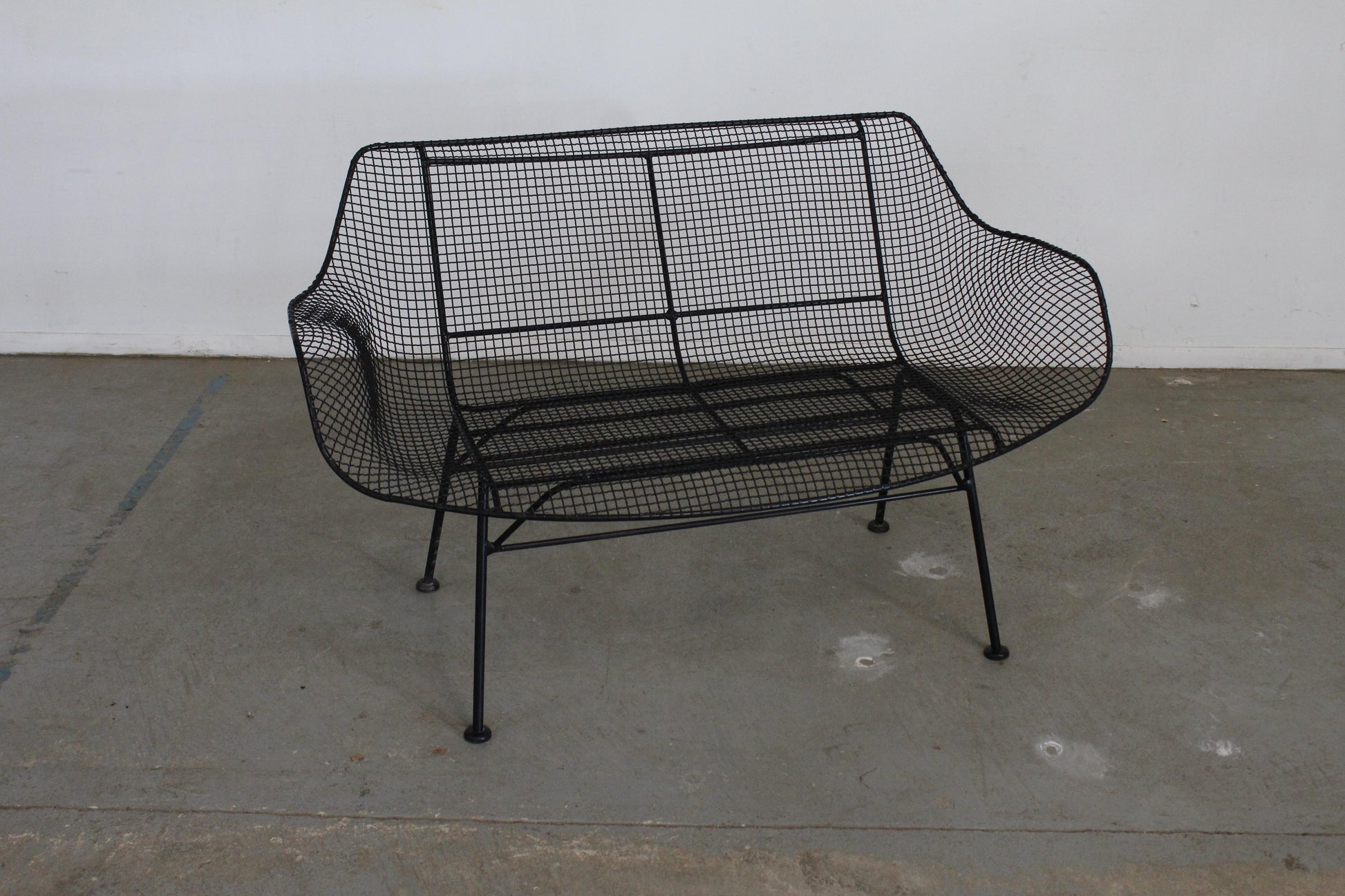 Black wire mesh bench on a concrete floor with a white wall background