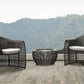 Two black wicker chairs with white cushions and a fire pit on a wooden deck with a mountainous background.