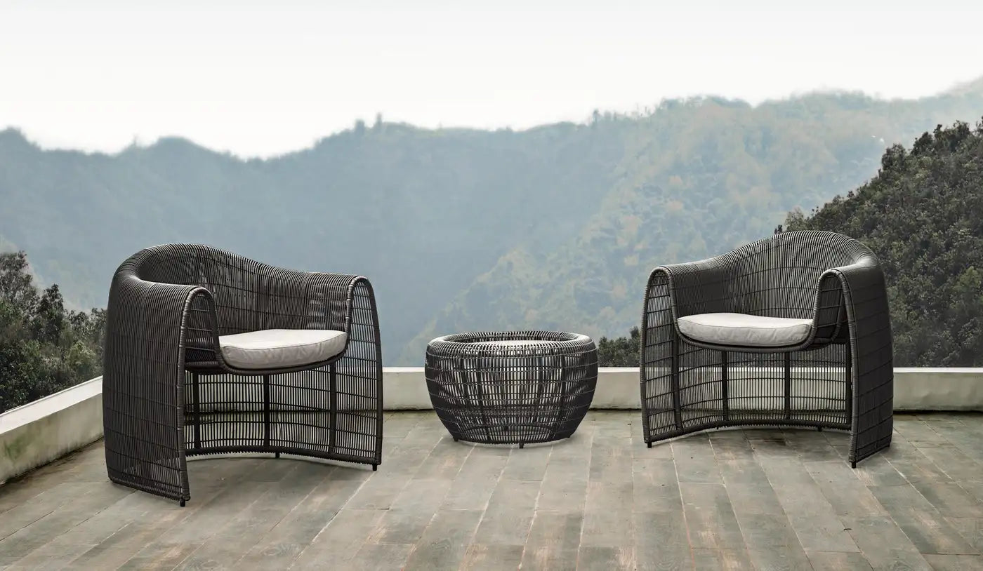 Two black wicker chairs with white cushions and a fire pit on a wooden deck with a mountainous background.