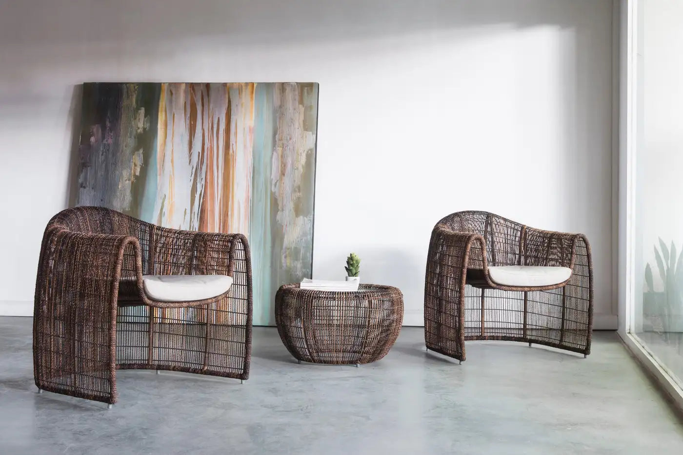 Two wicker chairs with a matching table in a modern room with abstract art on the wall.