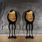 Two unique black and gold sculptures on a wooden floor with a textured wall in the background.