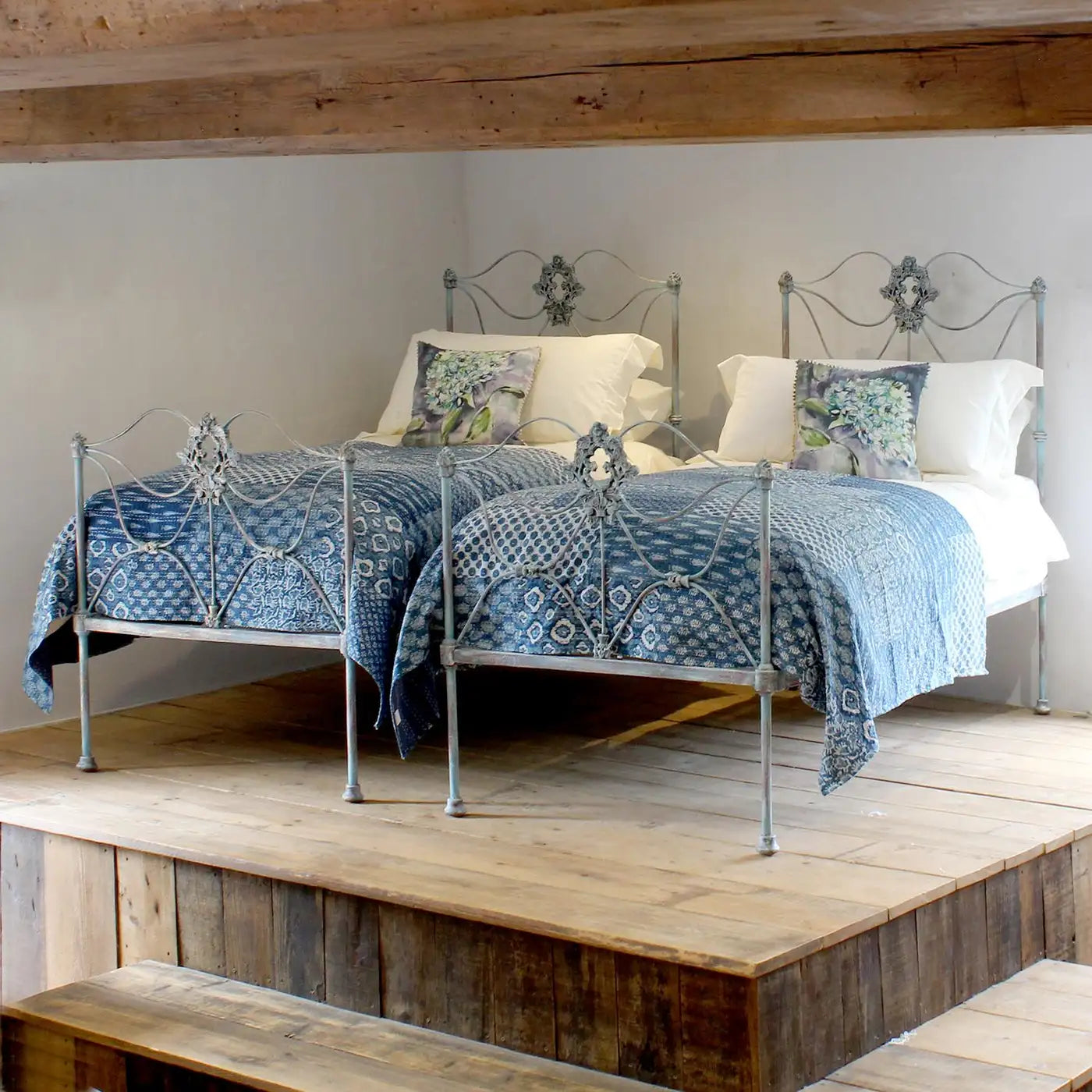 Pair of ornate metal beds with blue bedding in a room with wooden flooring and beams.