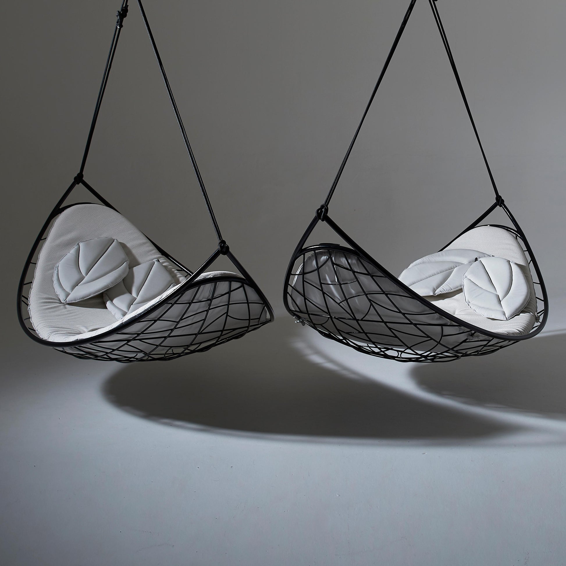 Two black mesh hanging chairs with white cushions on a gray background