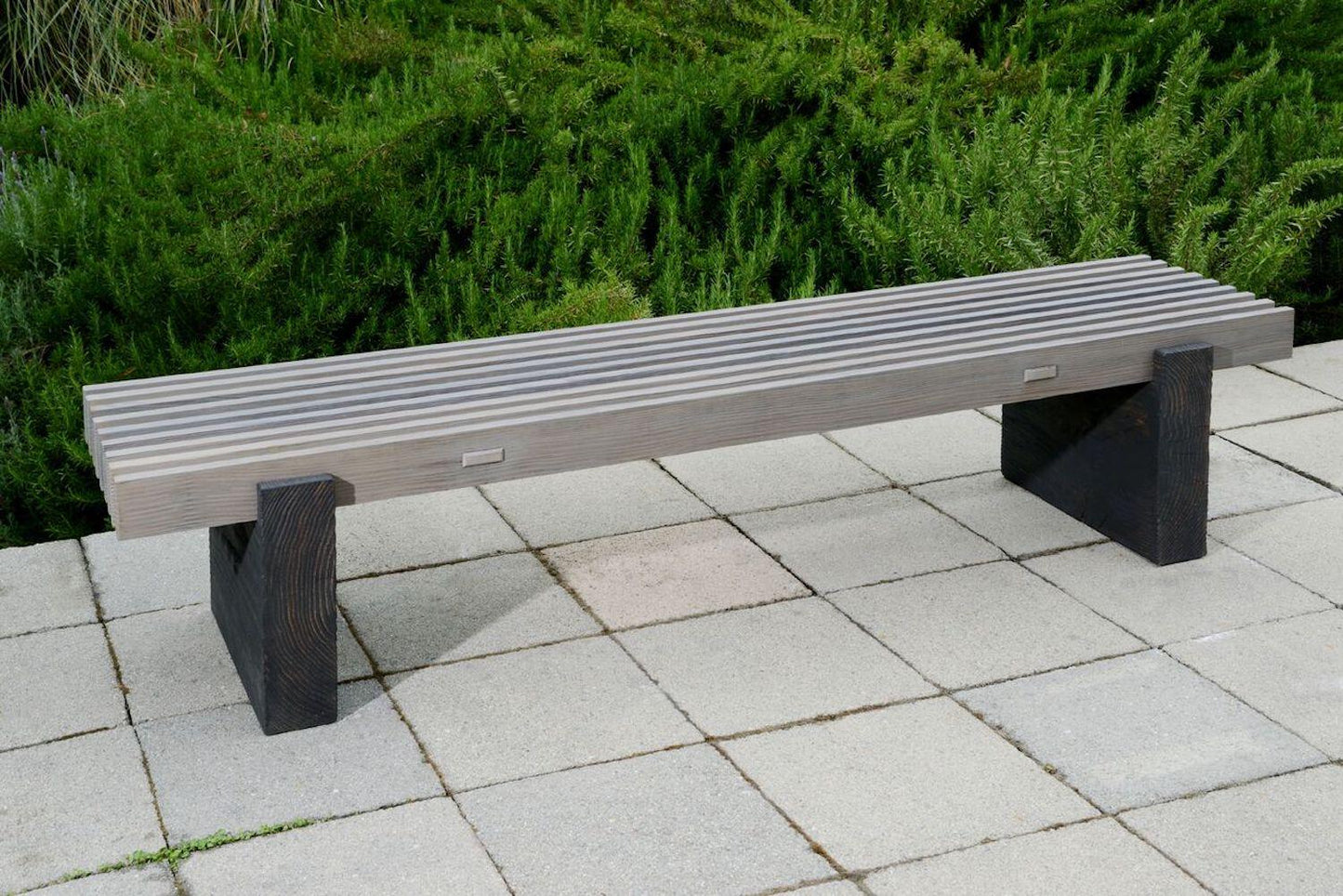Long wooden bench on a paved area with greenery in the background