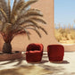 Two red chairs in a desert-like setting with a palm tree and beige wall.
