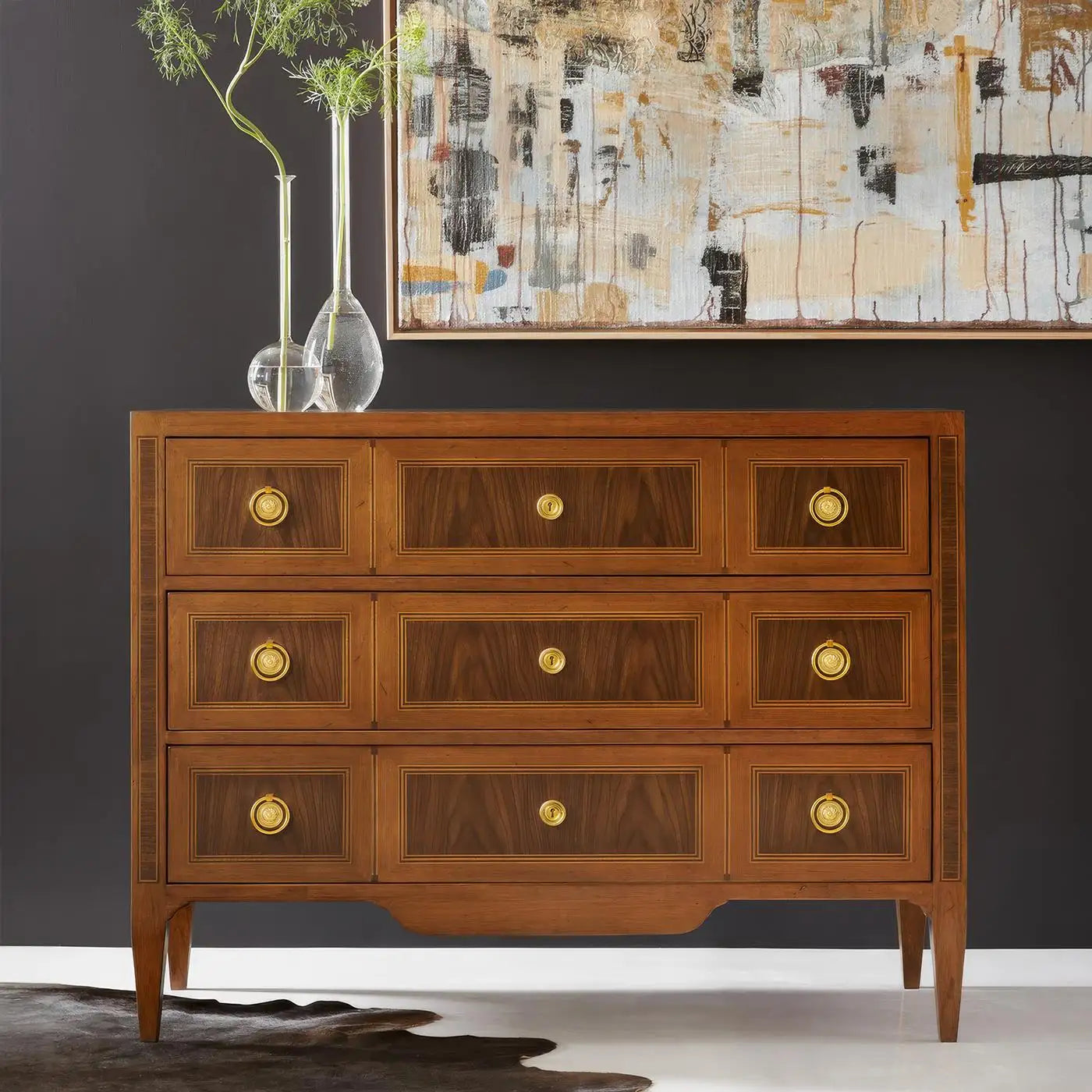 Wooden dresser with gold knobs against a dark wall with abstract art and vases.