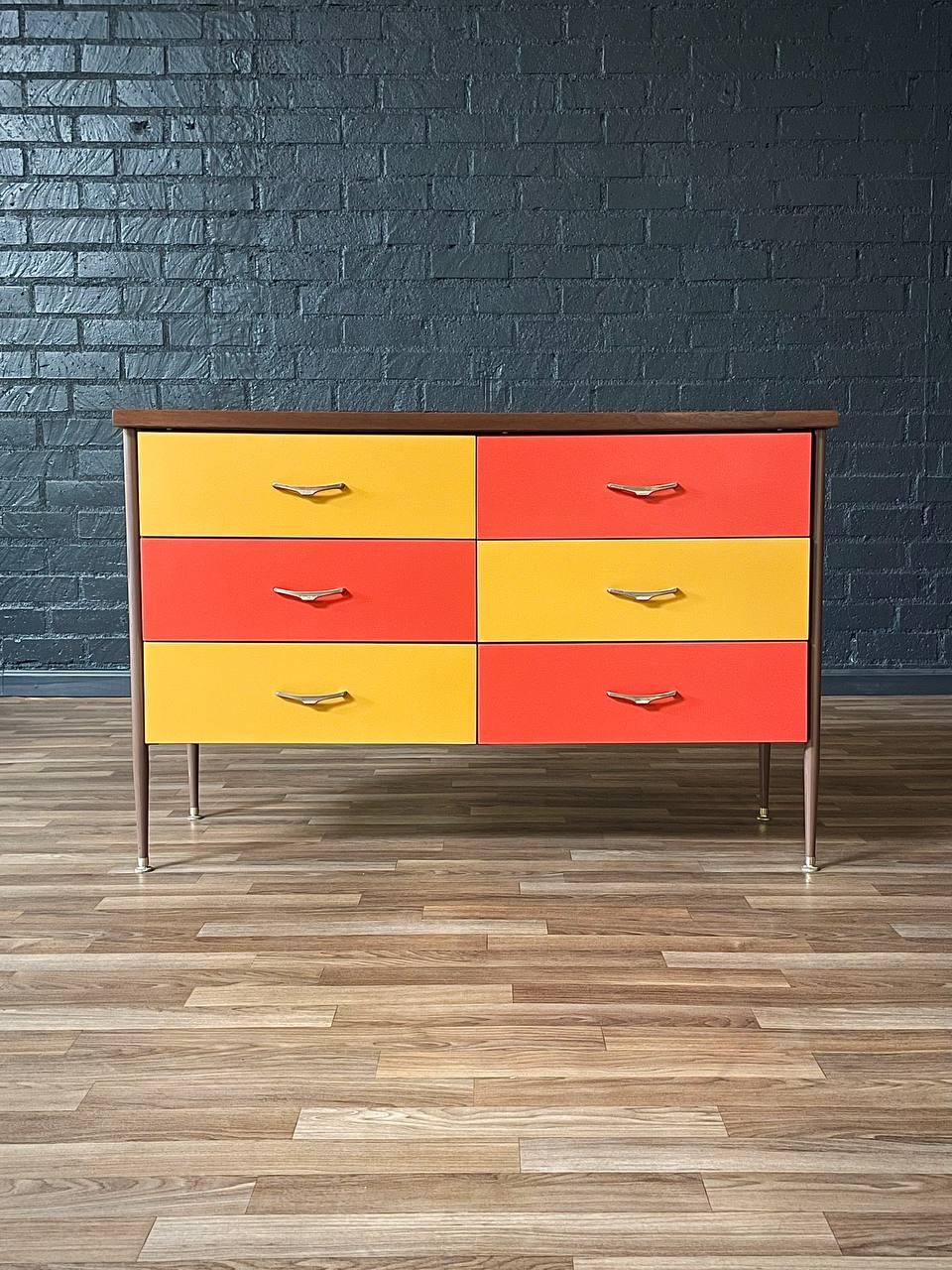 Colorful dresser with yellow and orange drawers against a blue brick wall.
