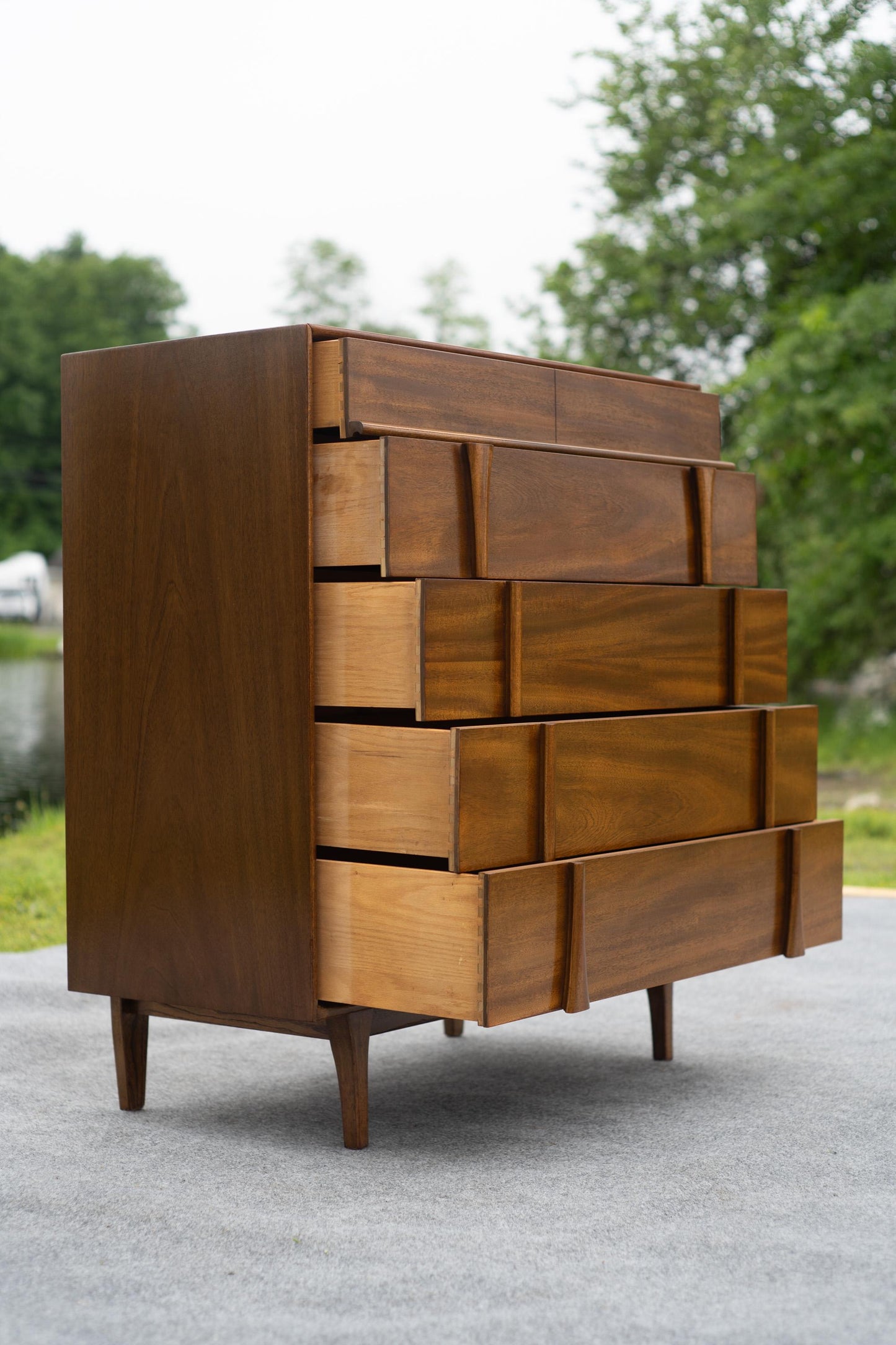 Wooden dresser with multiple drawers outdoors on a paved surface with greenery in the background